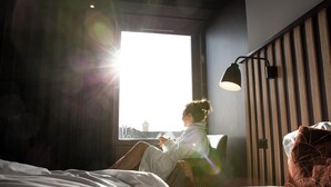 A room with a bed, a window with a view of a cityscape, and a wall-mounted lamp.