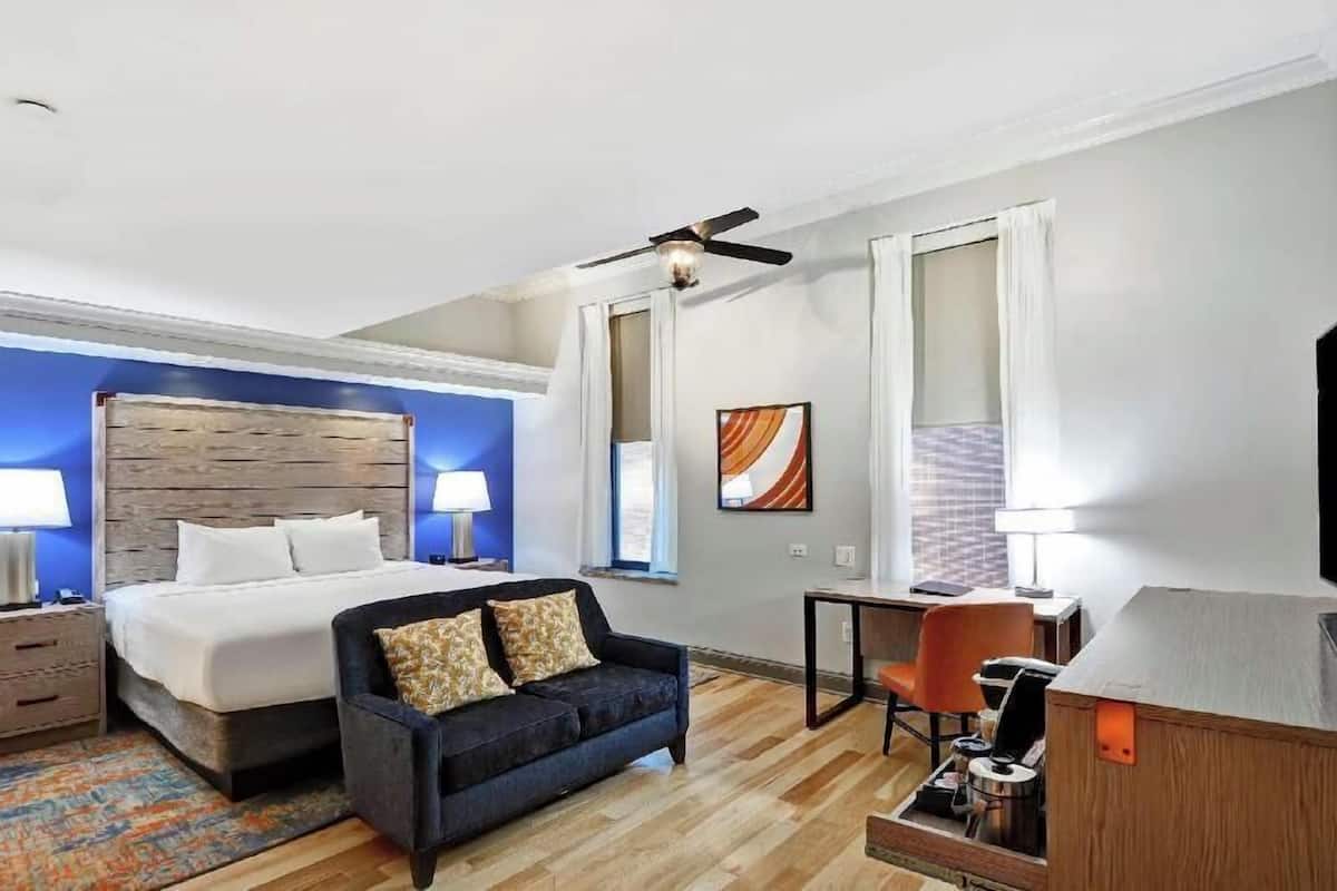 A hotel room with a bed, a sofa, a desk, a chair, a ceiling fan, and a TV.