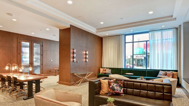 A modern hotel lobby with a wooden panel wall, a green sofa, a leather couch, a dining area with a table and chairs, and a large window with curtains. - The Cincinnatian Hotel Curio Collection by Hilton (Cincinnati)