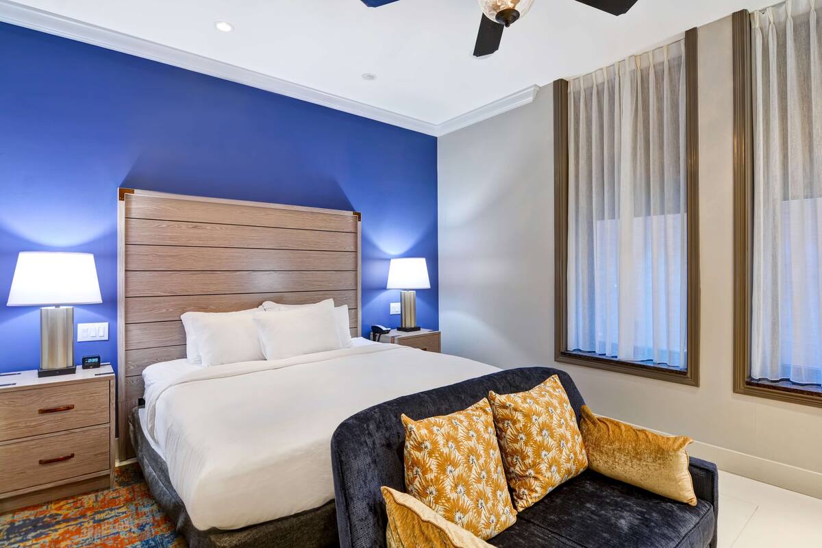 A hotel room with a large bed, two bedside tables with lamps, a dark sofa with cushions, a patterned rug, and a window with curtains.