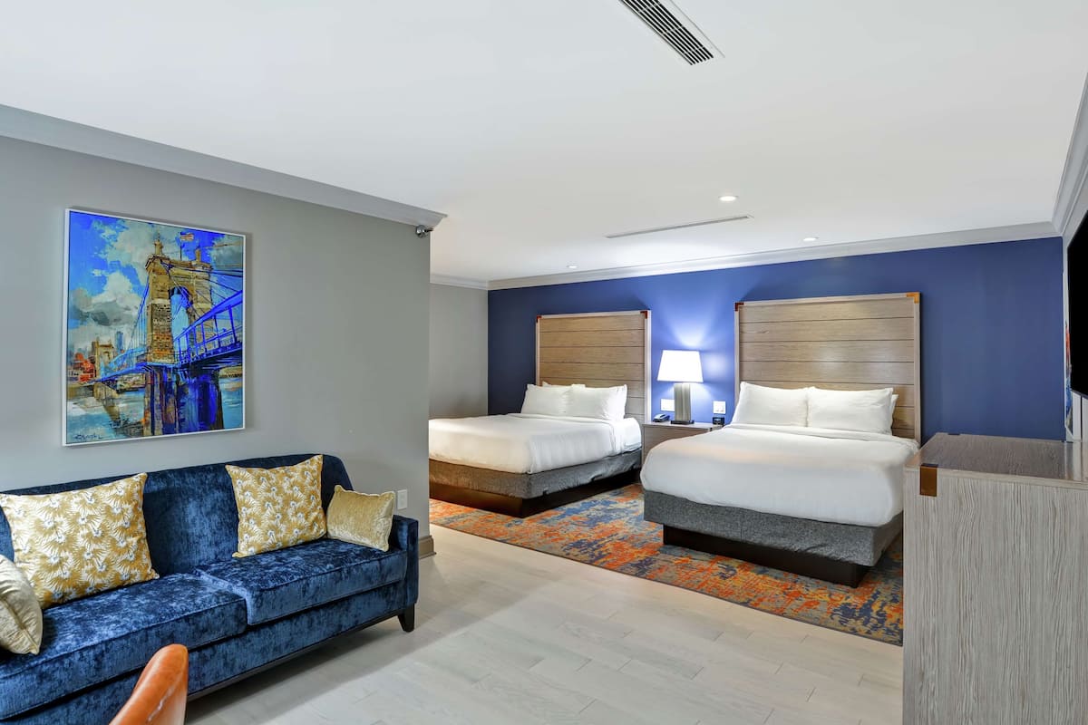 A hotel room with two beds, a blue sofa, and a painting of a bridge.