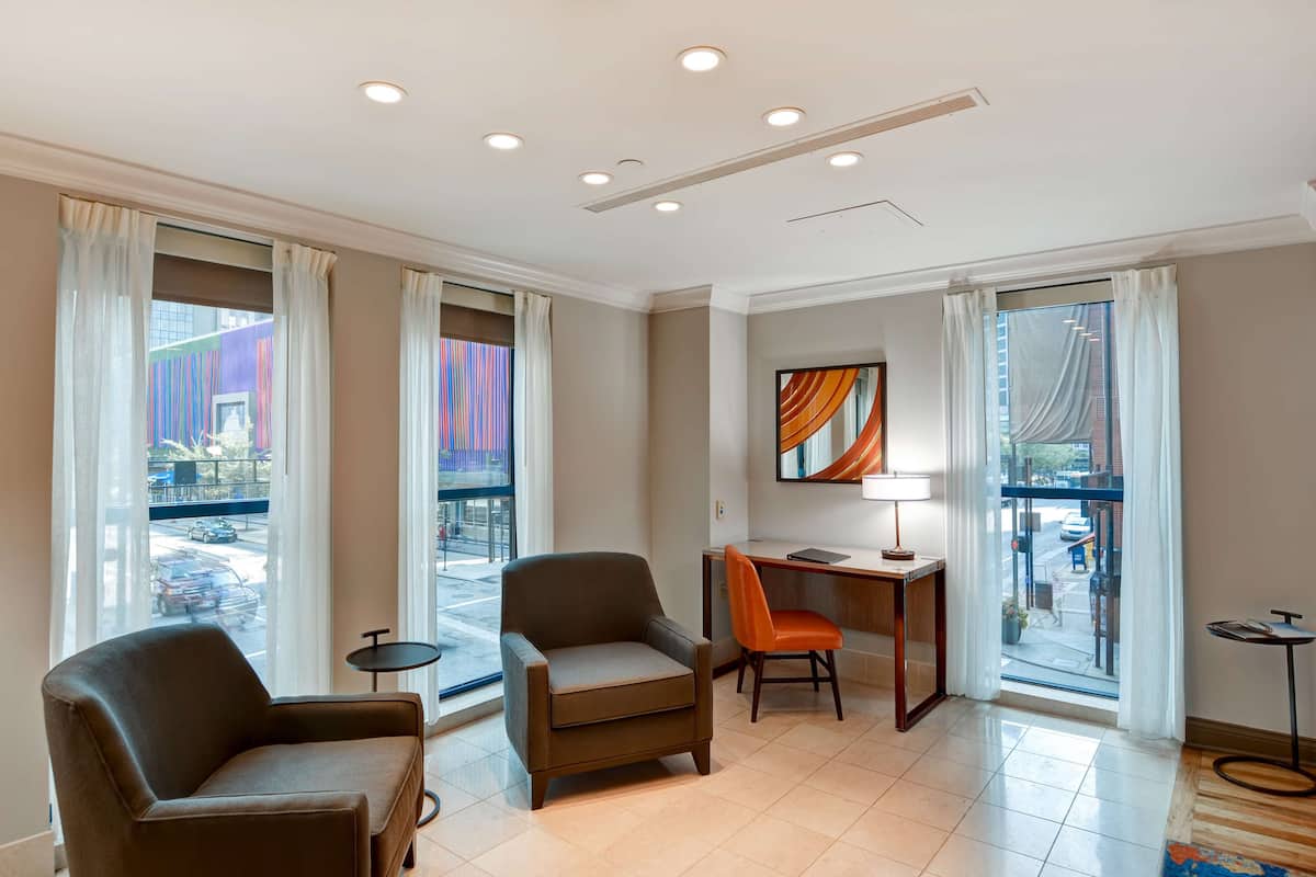 A modern hotel room with two armchairs, a desk, a small table, and a view of the street through large windows.