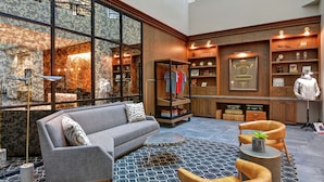 A spacious lobby with a gray sofa, a round coffee table, and a leather armchair. There is a large window with a view of a building outside, and a display of clothing and accessories on shelves. - The Cincinnatian Hotel Curio Collection by Hilton (Cincinnati)