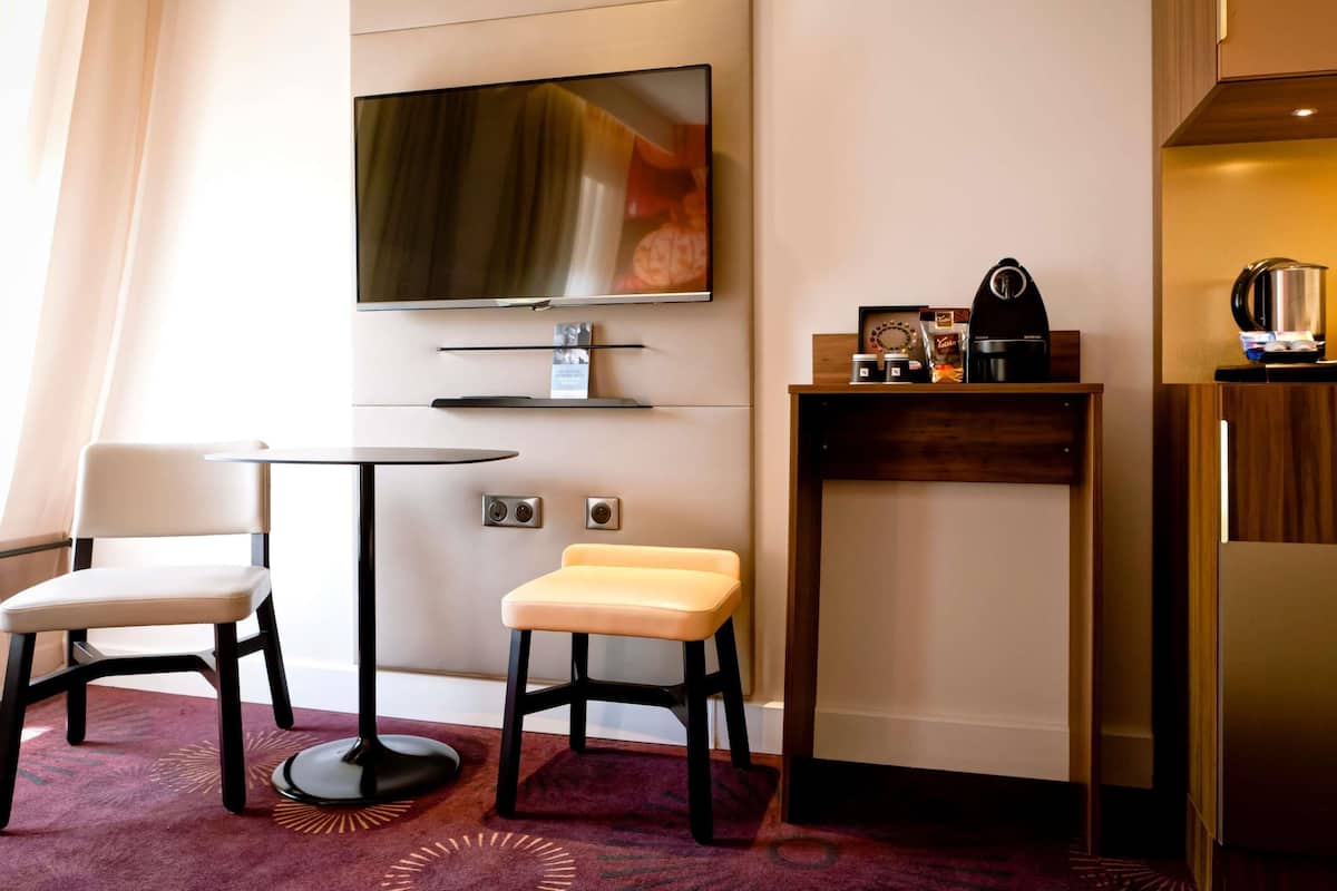 Quarto de hotel com televisão de tela plana na parede, uma mesa pequena com cadeira, um banquinho, máquina de café e geladeira.