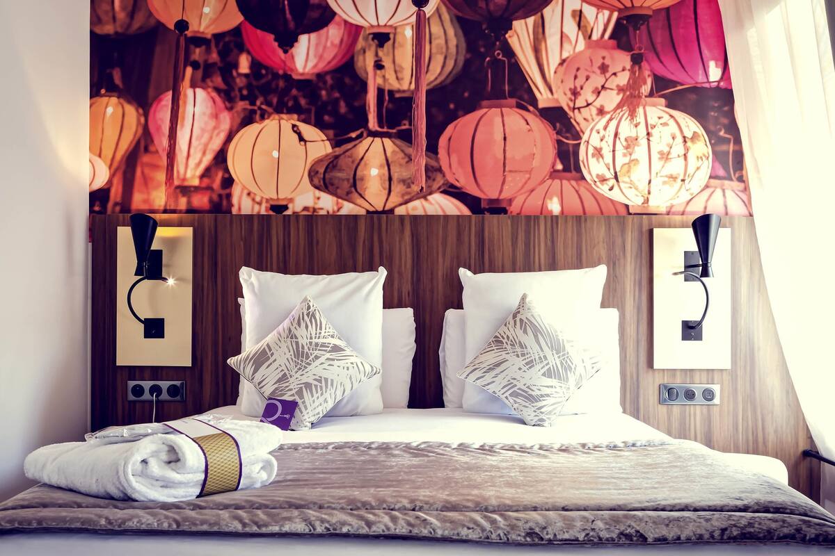 Ein Hotelzimmer mit einem Holzkopfteil, zwei weißen Kissen, einem gefalteten Handtuch und einer Wand mit hängenden Laternen.