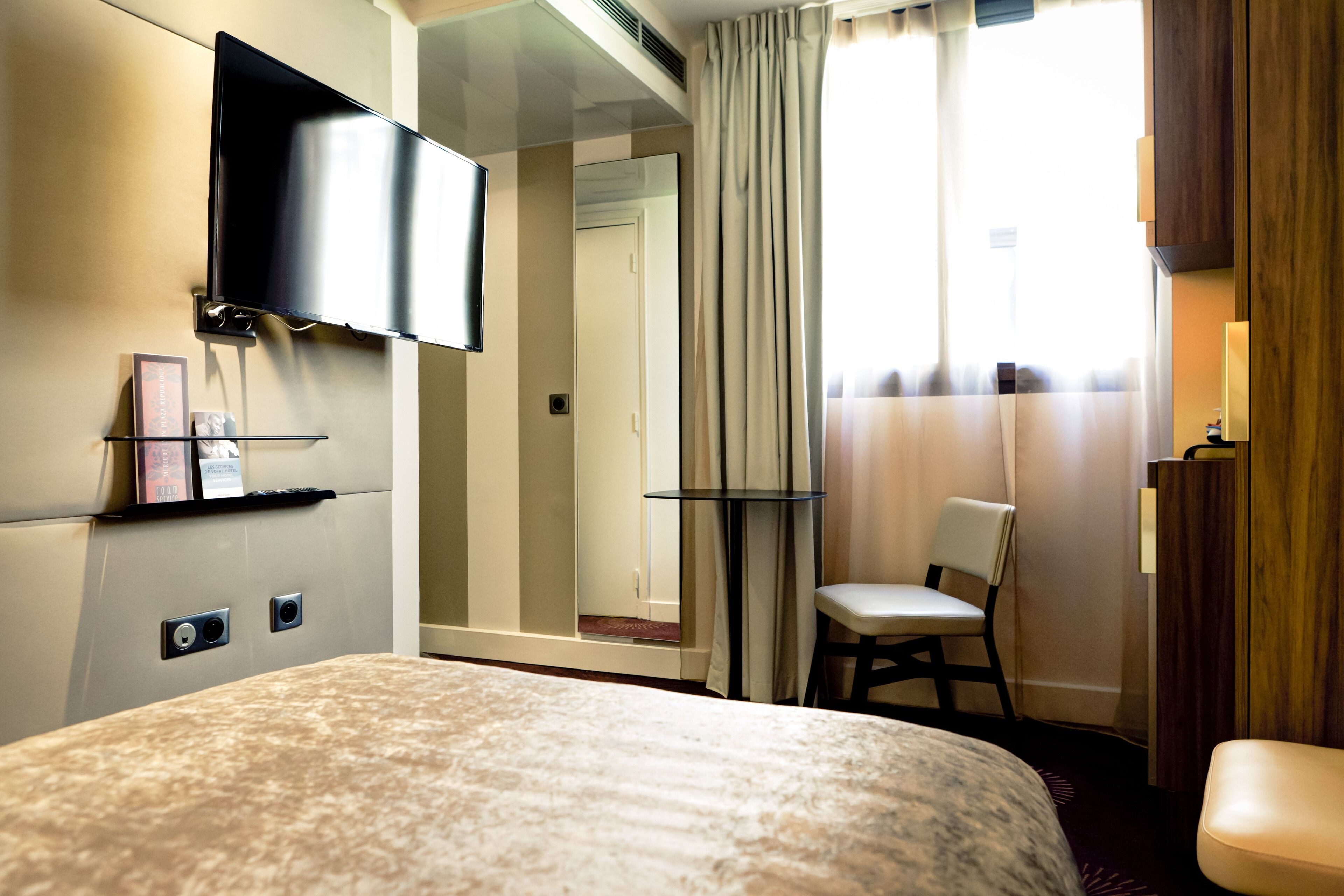 A hotel room with a large bed, a television mounted on the wall, a small table, a chair, and a mirror.
