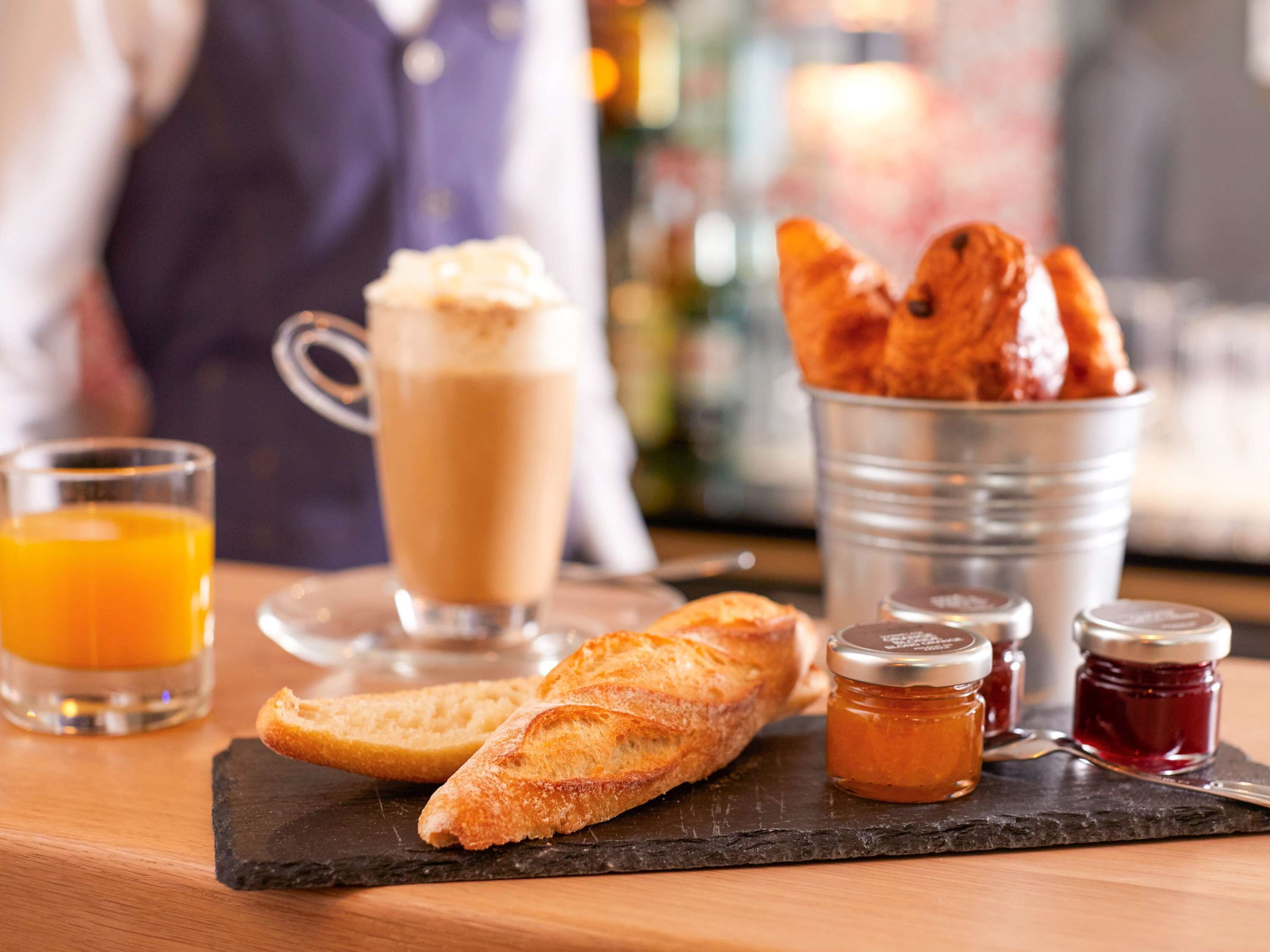 A breakfast setting with a glass of orange juice, a cup of coffee, a baguette, croissants, and two jars of jam on a slate board.