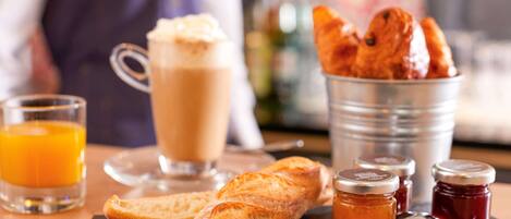 A breakfast setting with a glass of orange juice, a cup of coffee, a baguette, croissants, and two jars of jam on a slate board.