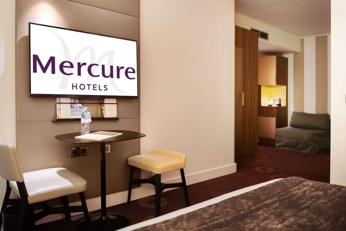 Eine Hotellobby mit einer Rezeption, einer Sitzecke mit Stühlen, einem großen Schild für Mercure Hotels und einem Blick auf ein Zimmer mit Bett und Fenster.