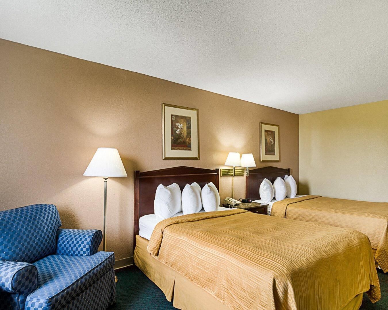 A hotel room with two beds, a chair, a lamp, and two framed pictures on the wall.