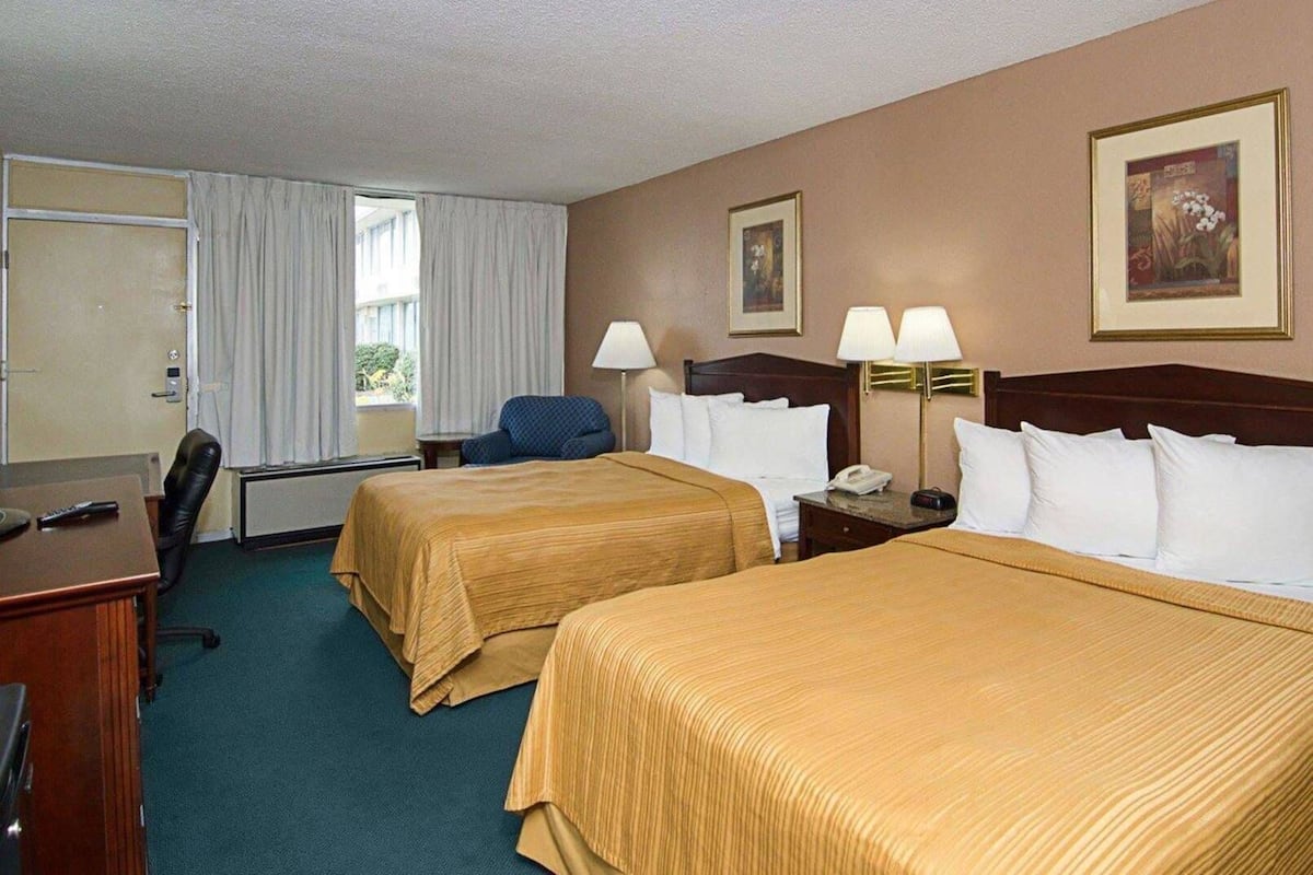 A hotel room with two beds, a desk, a chair, a lamp, and a painting on the wall.