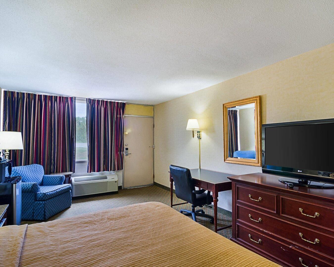 Ein Hotelzimmer mit Bett, Schreibtisch, Stuhl, Fernseher und einem Fenster mit Vorhängen.