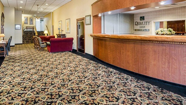 A hotel reception area with a patterned carpet, a reception desk, and a staircase leading to another floor. - Quality Inn Shenandoah Valley (New Market)