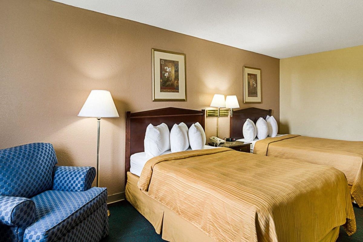 A hotel room with two beds, a chair, a nightstand, and two lamps.
