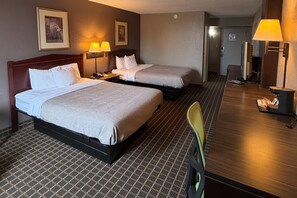A hotel room with two beds, a desk, a chair, and a lamp.