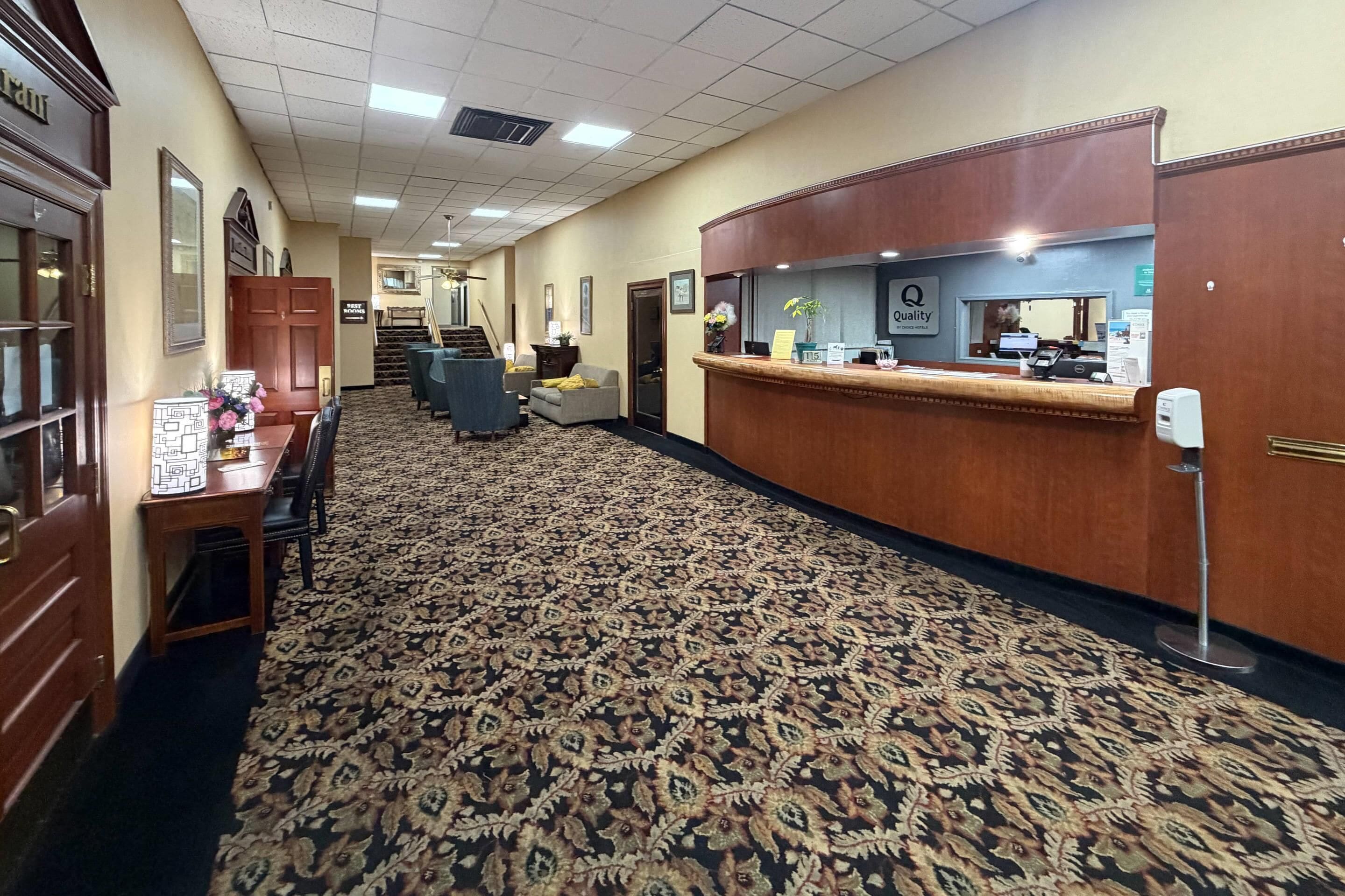 A hotel lobby with a reception desk, a staircase, and a seating area with a patterned carpet.