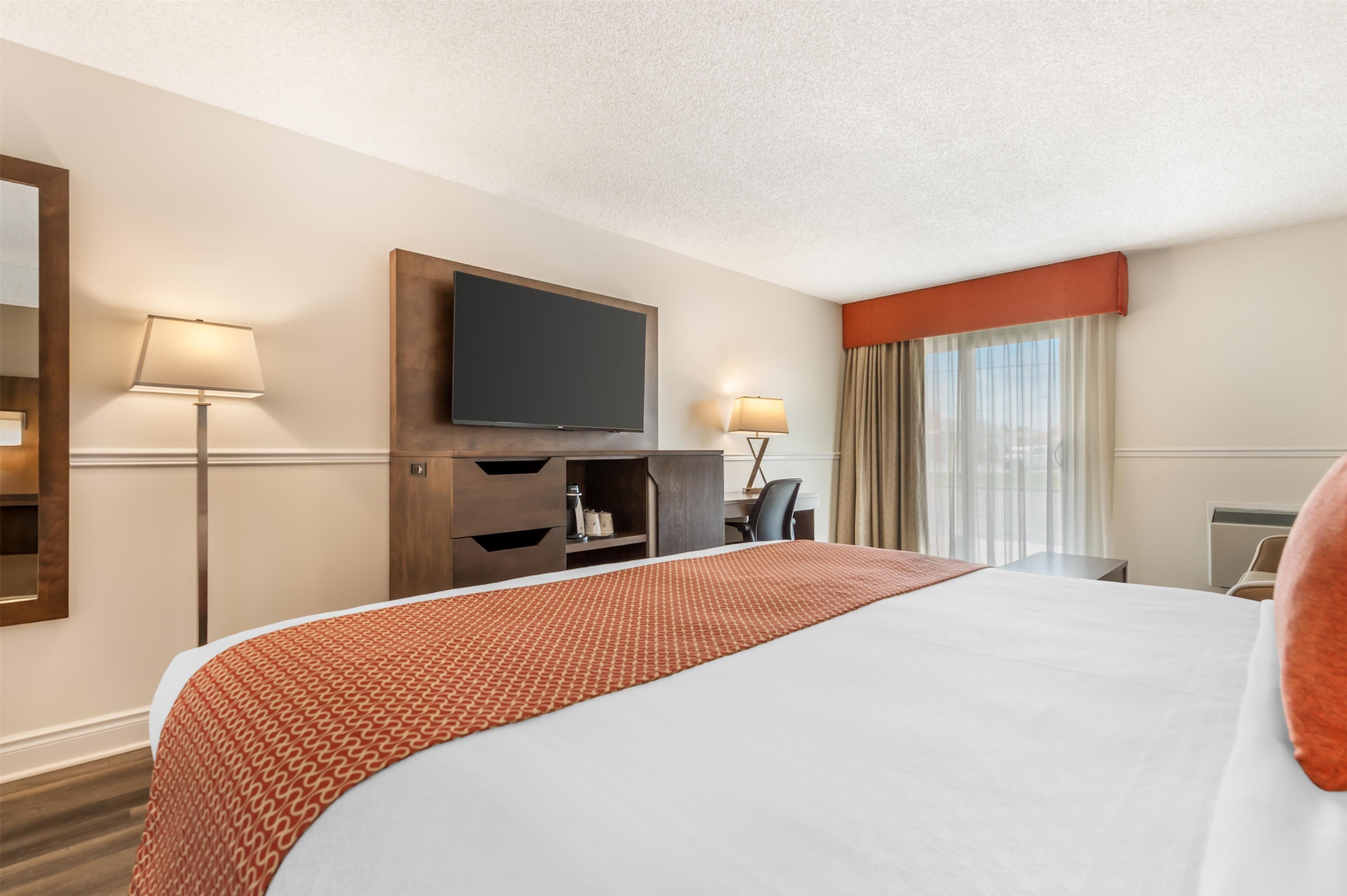 A hotel room with a large bed, a flat-screen TV mounted on the wall, a desk with a chair, and a window with curtains.