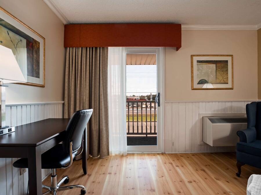 A hotel room with a wooden floor, a desk with a chair, a bed, a window with curtains, and a radiator.