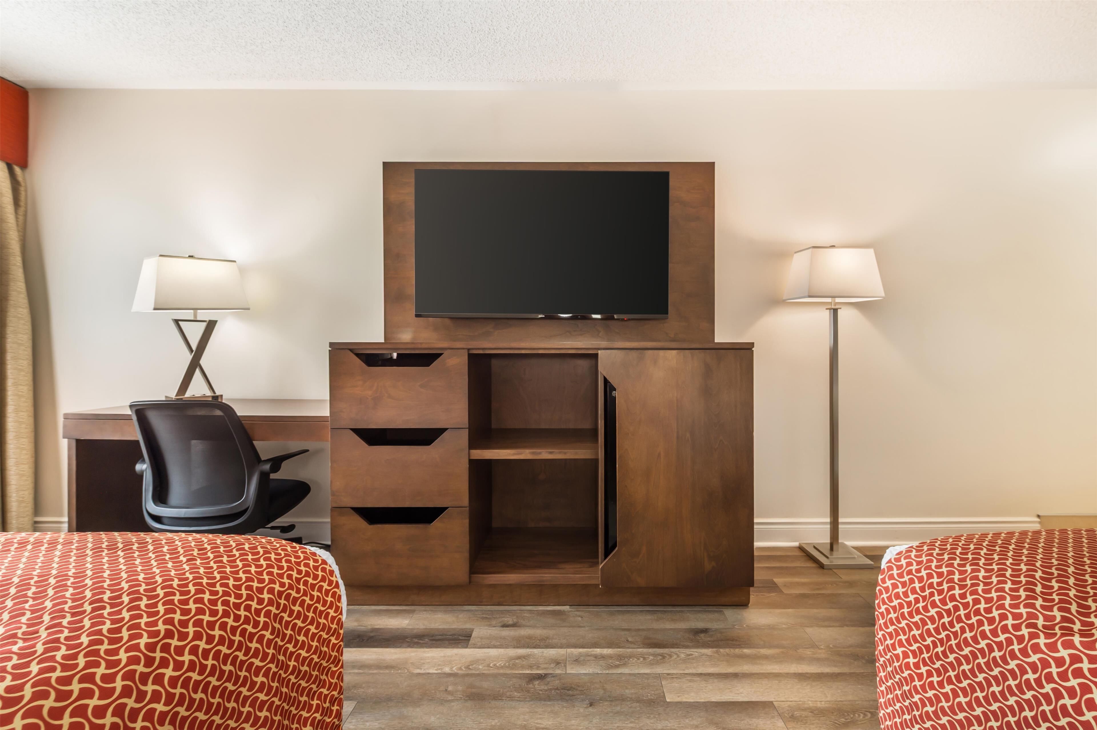 A hotel room with a TV mounted on a wooden cabinet, a desk with a chair, two beds with patterned bedding, and two floor lamps.