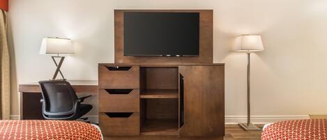 Una habitación de hotel con televisor montado en un mueble de madera, un escritorio con silla, dos camas con ropa de cama estampada y dos lámparas de pie.