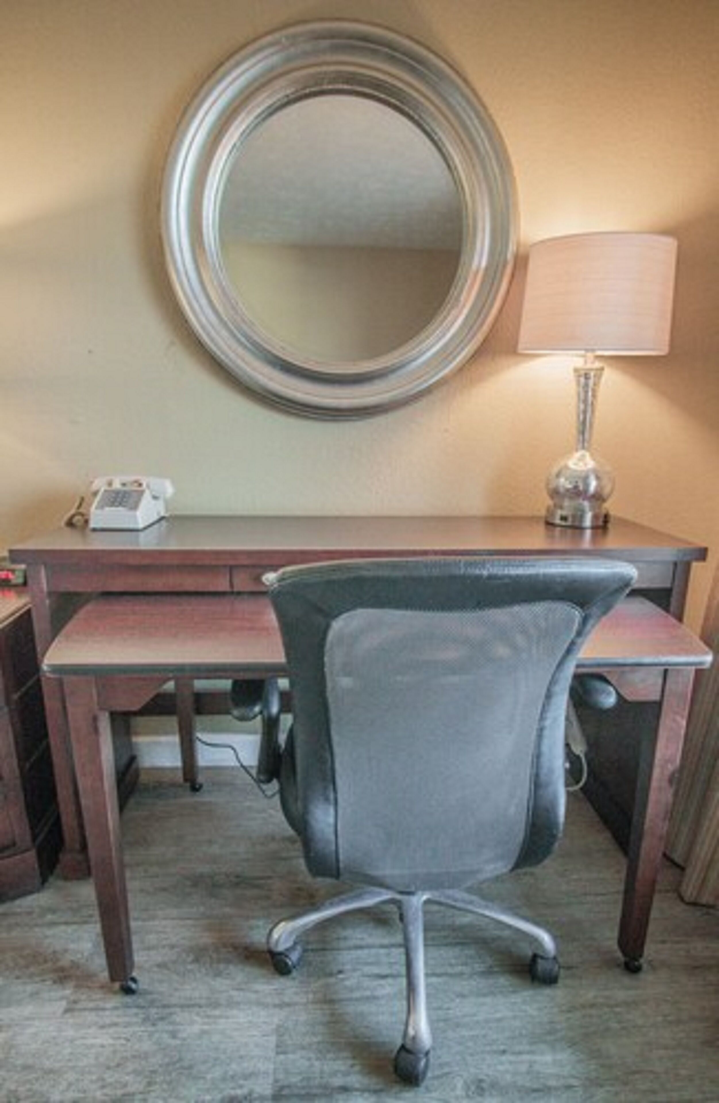 Een hotelkamer met een houten bureau, een grijze bureaustoel, een ronde spiegel, een lamp en een telefoon.