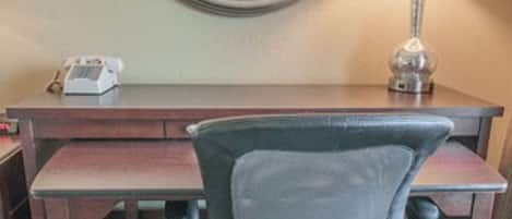 Een hotelkamer met een houten bureau, een grijze bureaustoel, een ronde spiegel, een lamp en een telefoon.