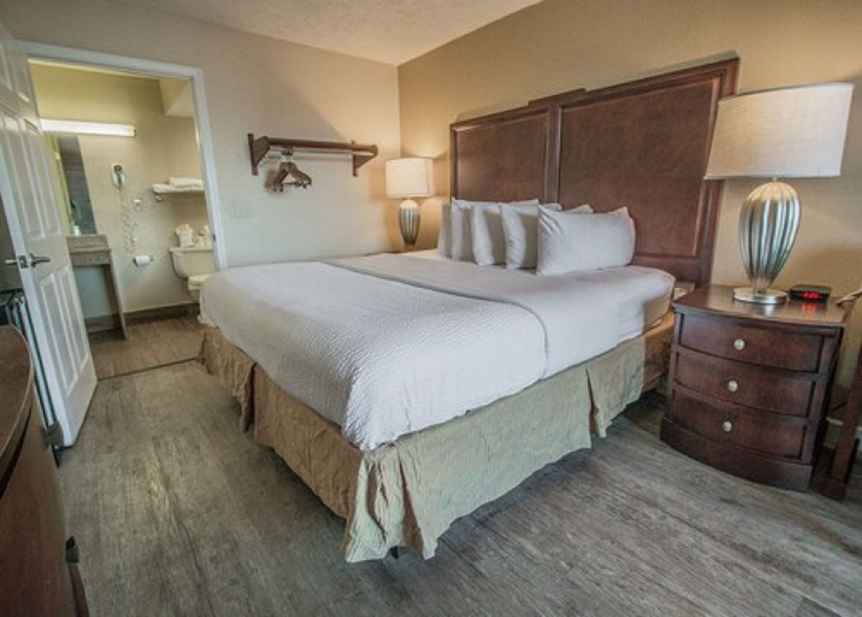 Una habitación de hotel con una cama grande, cabecera de madera, mesita de noche con lámpara y puerta que da a un baño.