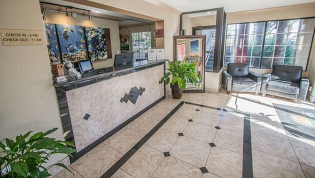 Ein Hotel-Rezeptionsbereich mit Marmor-Theke, Schild für Check-in/Check-out, Empfangstresen, Zimmerpflanze und einer Sitzecke mit Stühlen.