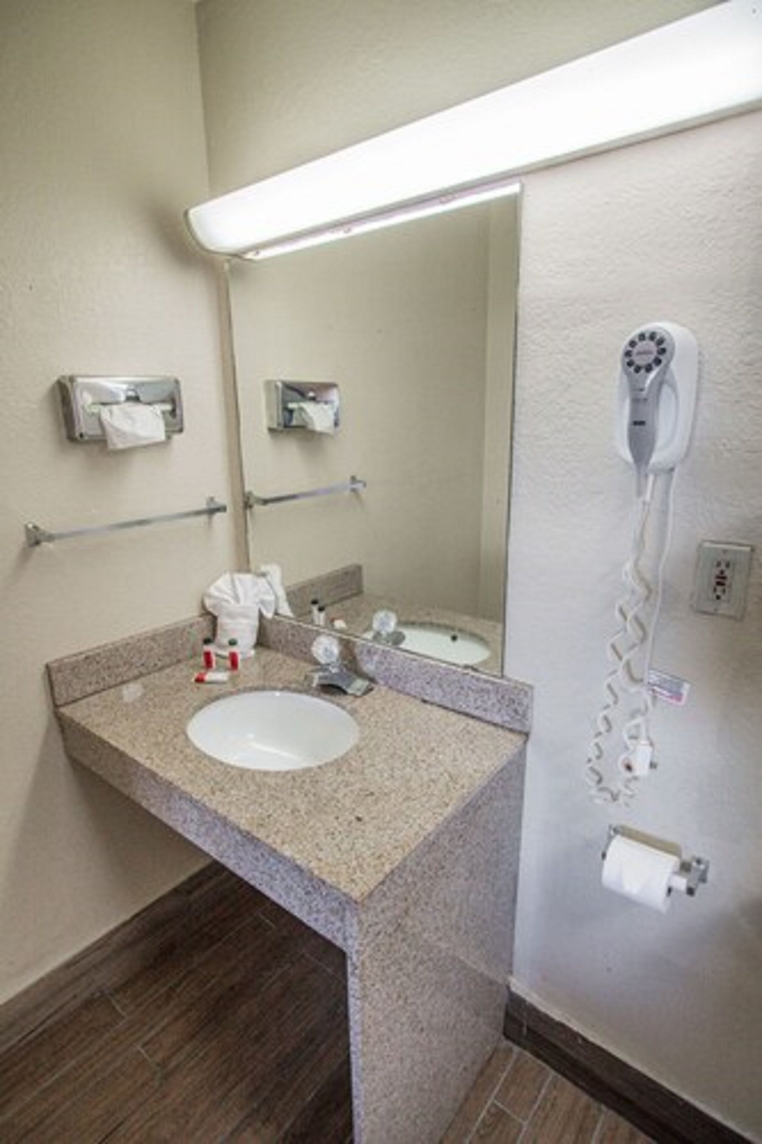Un baño con lavamanos de encimera de granito, un espejo, un secador de pelo montado en la pared y un dispensador de toallas.