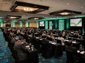 A conference room with rows of tables and chairs, a stage with a speaker, and a large screen displaying a presentation. - Wyndham Garden Oklahoma City Conference Ctr/Airpt/Bricktown (Oklahoma City)