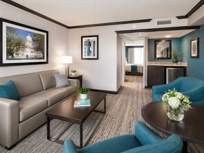 A hotel room with a sofa, a coffee table, a dining table with a vase of flowers, a kitchenette, and a bathroom visible through an open door.