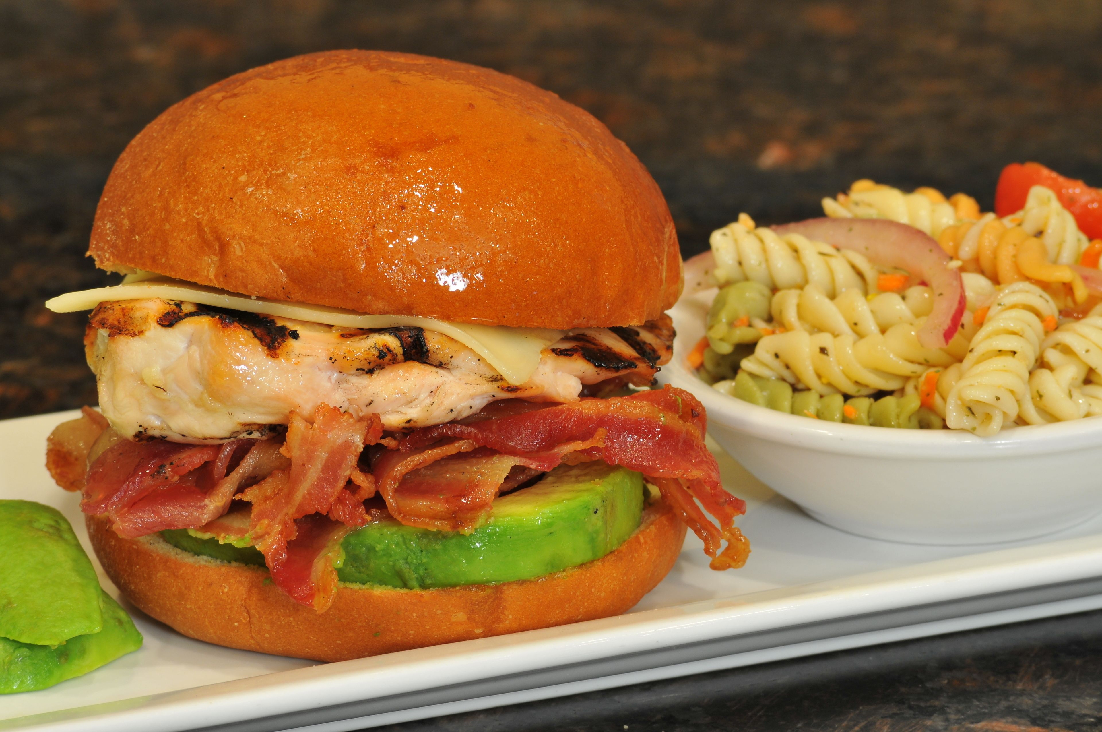 A sandwich with grilled chicken, bacon, and avocado on a bun, alongside a bowl of pasta salad.