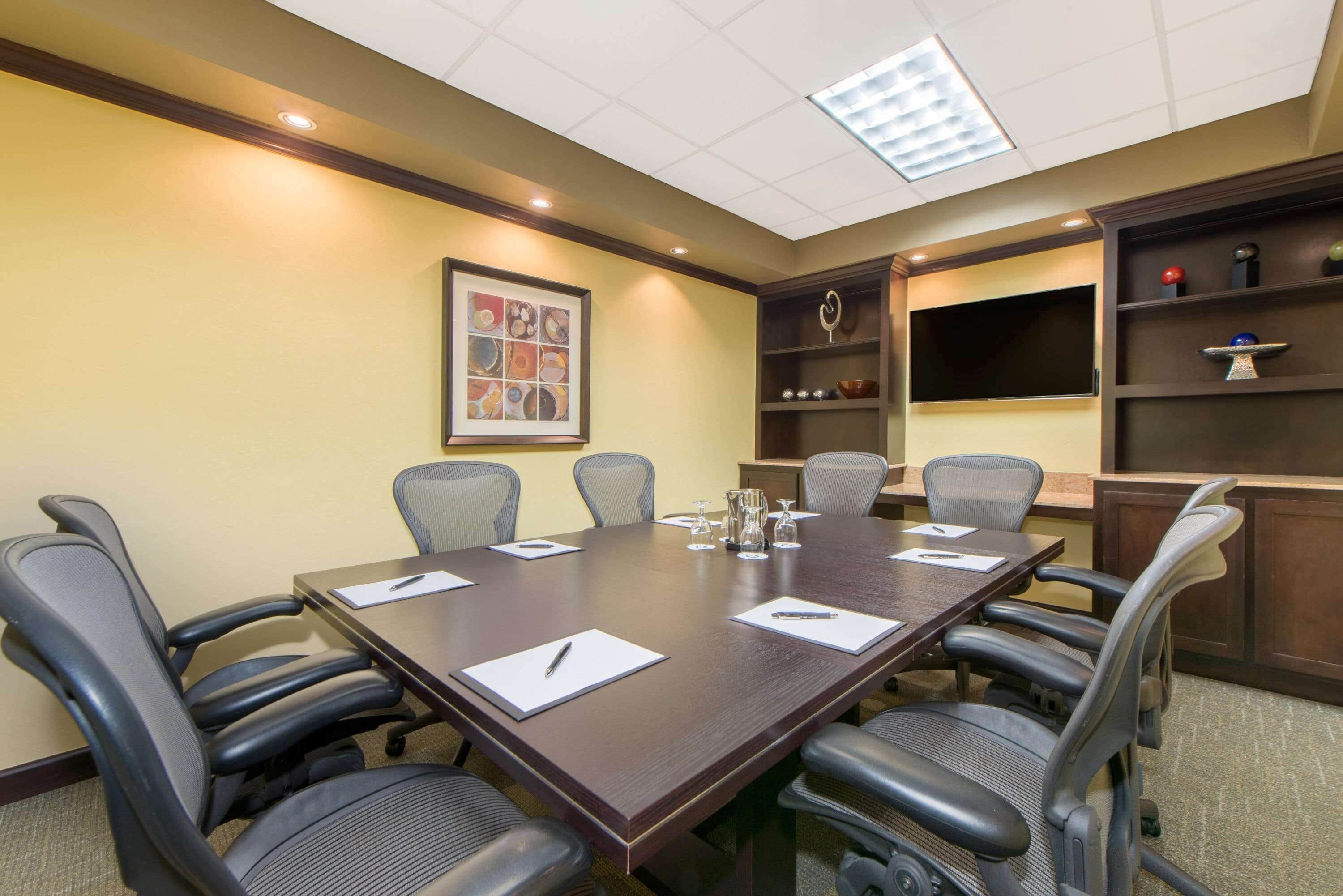 A conference room with a long table, six chairs, and a television on the wall.