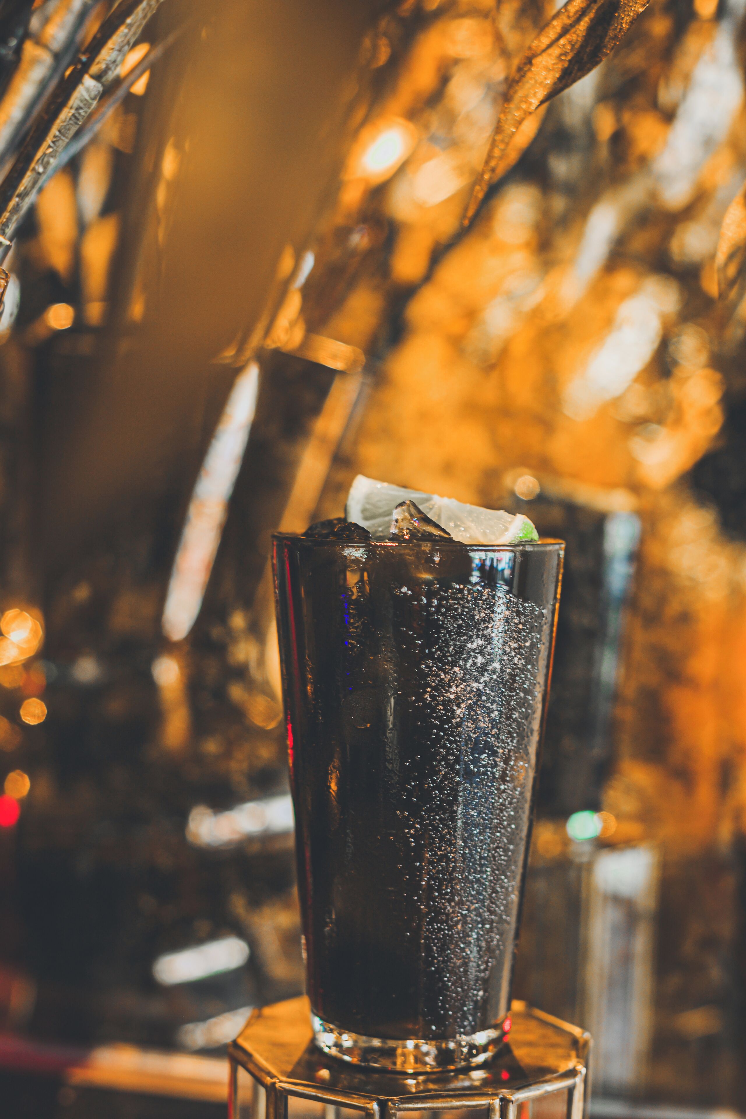 A tall glass with a dark beverage, ice, and a lime wedge, set against a blurred background with warm lighting.