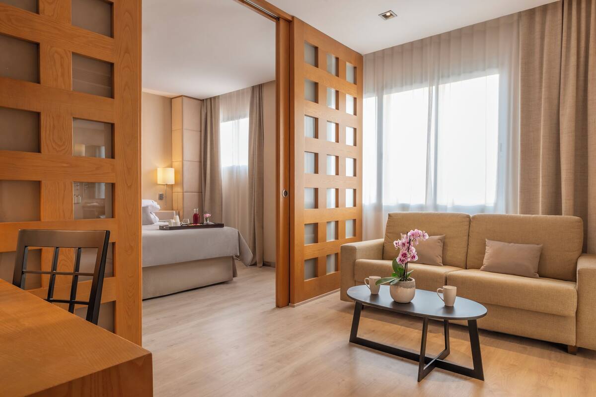 A hotel room with a wooden partition, a sofa, a bed, a dining table, and a coffee table with a flower vase.