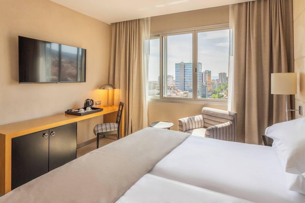 A hotel room with a bed, a desk, a chair, a TV, and a window with a city view.