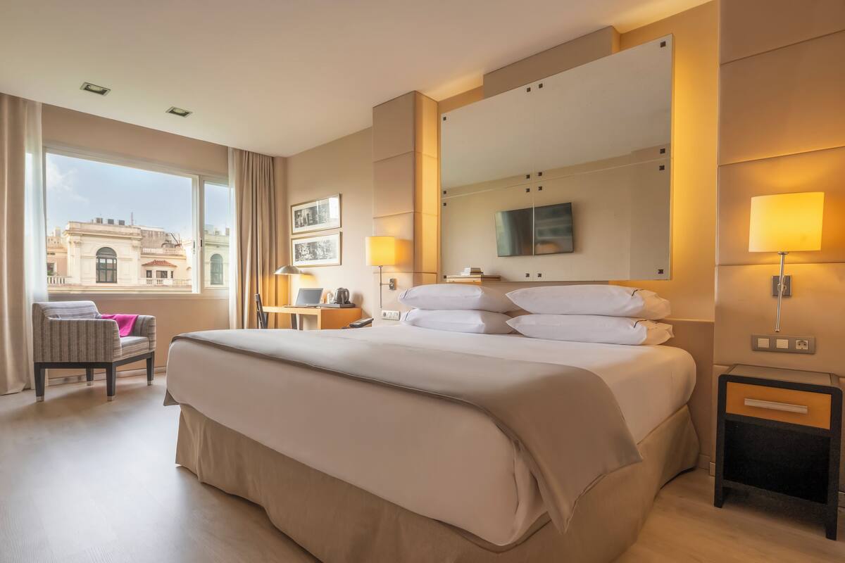 A hotel room with a large bed, a desk with a lamp, a chair, and a view of the cityscape.