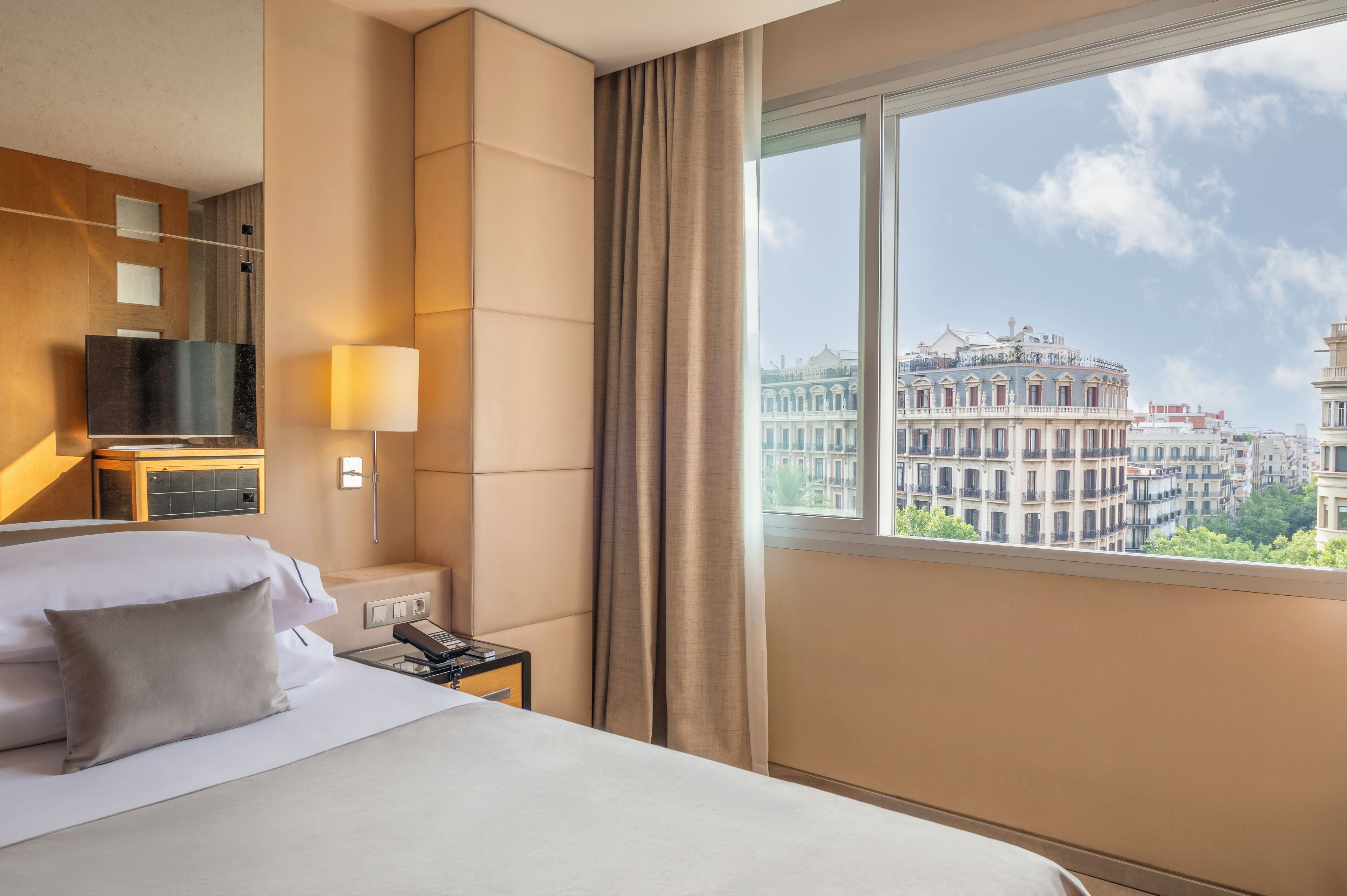 A hotel room with a bed, bedside table, lamp, and a view of a historic building through the window.