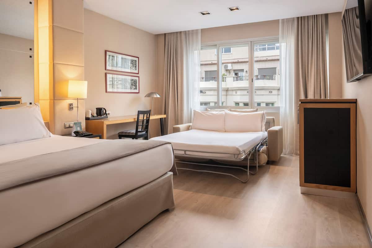 A hotel room with a large bed, a desk with a chair, a small table with a lamp, and a TV.