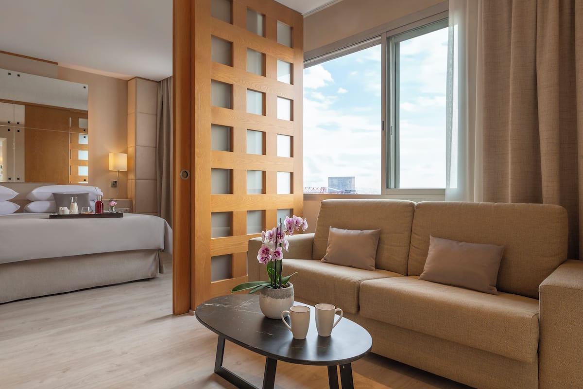 A hotel room with a bed, a sofa, a coffee table with a flower vase, and a window with curtains.