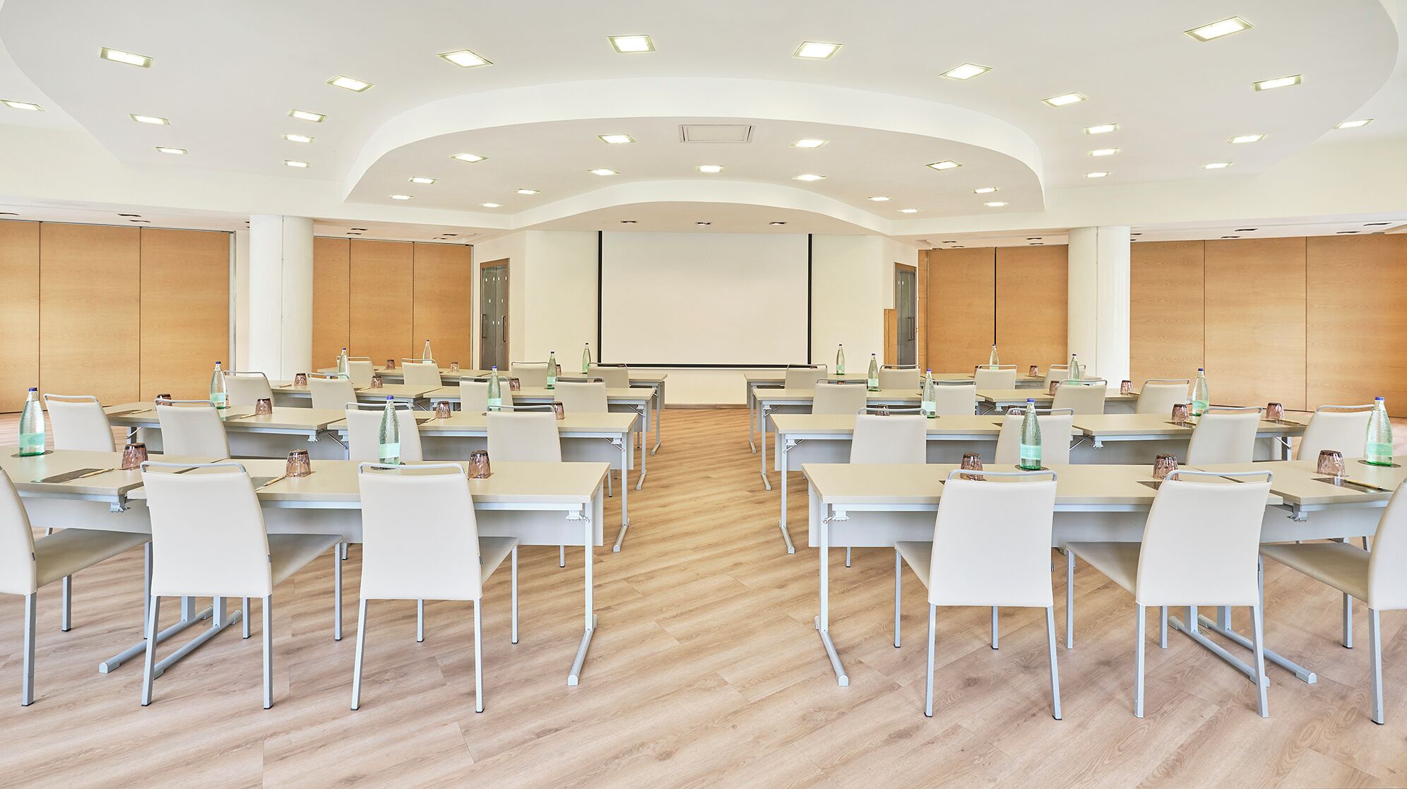 Un salón de conferencias con varias mesas y sillas, equipado con botellas de agua y vasos, y una pantalla grande al frente.
