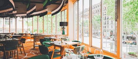 A restaurant with large windows, green chairs, and tables set with wine glasses and cutlery.