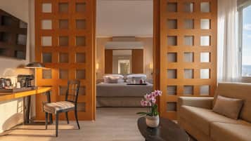 Quarto de hotel com cama, sofá, área de refeições com mesa e cadeiras, e vista através de um painel de madeira.