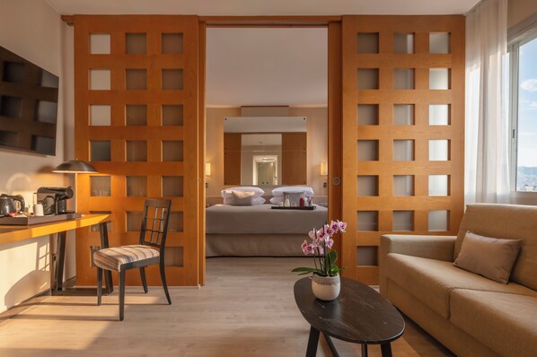 Habitación de hotel con cama, sofá, zona de comedor con mesa y sillas, y vistas a través de una pared divisoria de madera.