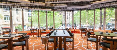 Un restaurante con mesas y sillas dispuestas en un ambiente amplio, ventanas grandes que ofrecen vistas de la ciudad y una alfombra anaranjada con diseño.