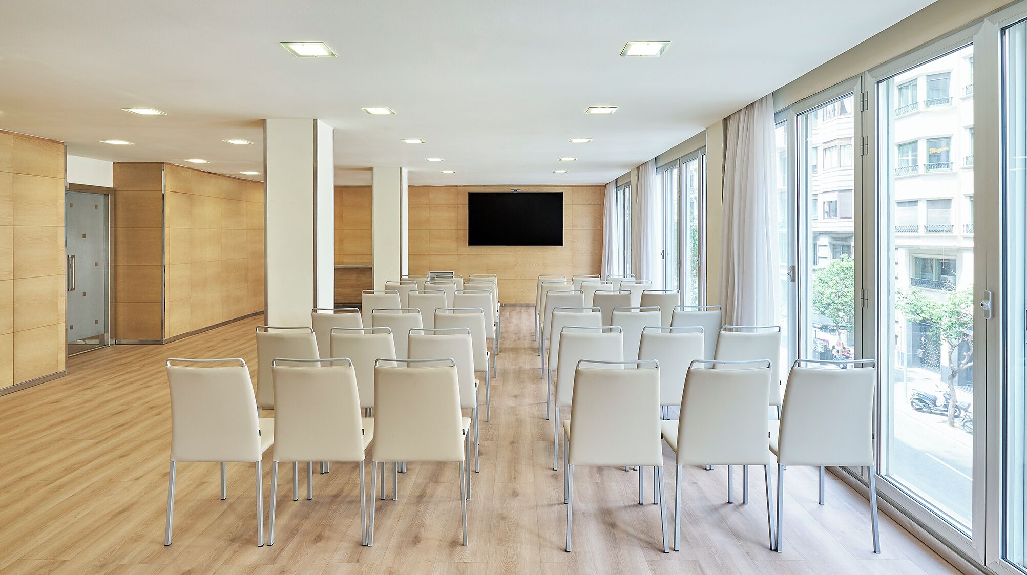 A conference room with rows of white chairs, a large flat-screen TV on the wall, and large windows offering a view of the street.