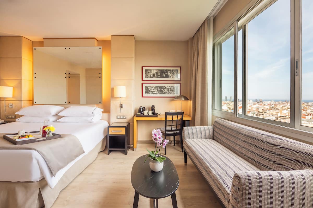 A hotel room with a bed, a desk, a chair, a sofa, a small table with flowers, and a large window with a city view.