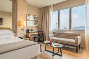 Ett hotellrum med en säng, ett skrivbord, en stol, en soffa, ett litet bord med en drink och frukt på, och ett fönster med utsikt över staden.