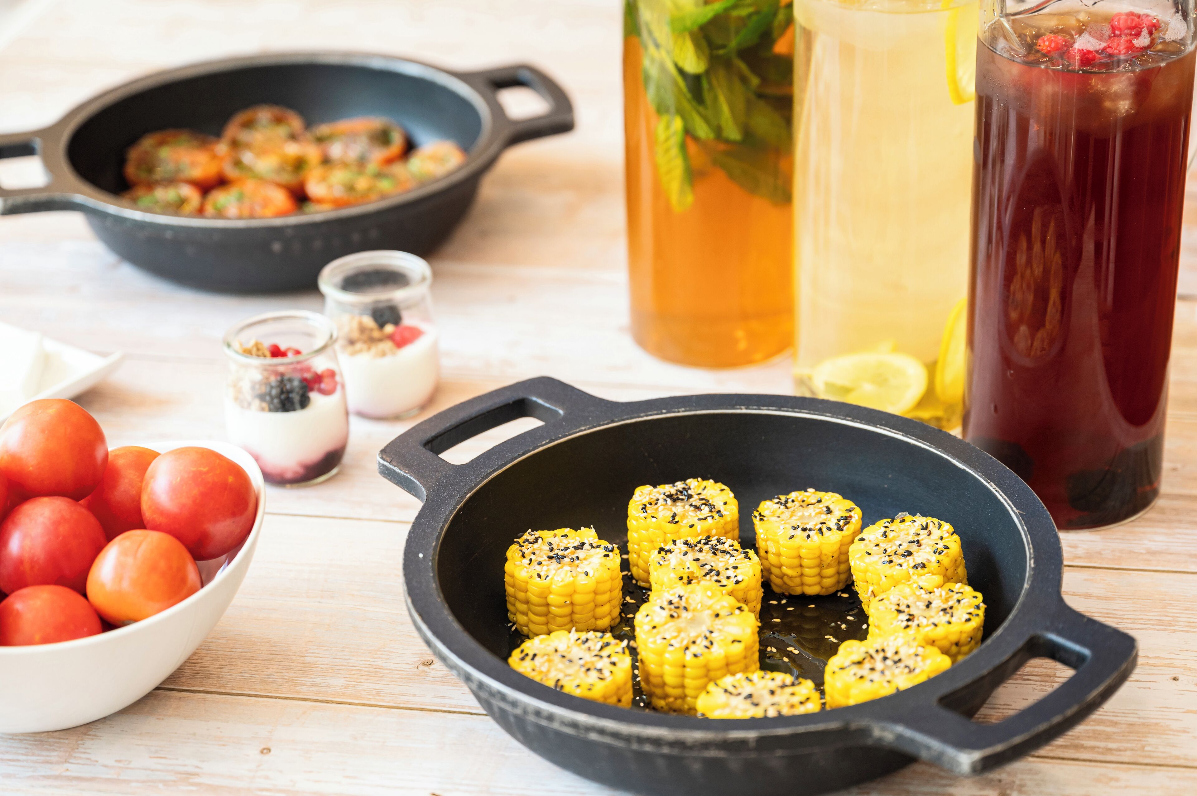 A kitchen with a skillet containing corn on the cob, a bowl of tomatoes, and a pitcher of iced tea.