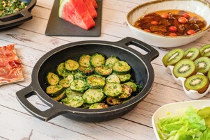 A skillet with sliced zucchini garnished with herbs, a bowl of red fruit, and a plate of sliced kiwi.